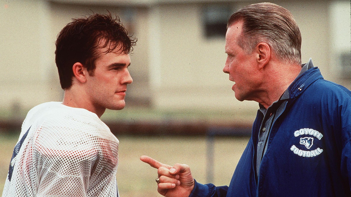 James Van Der Beek in a white football uniform stares at Jon Voight in a blue jacket, pointing his finger in a scene from "Varsity Blues"