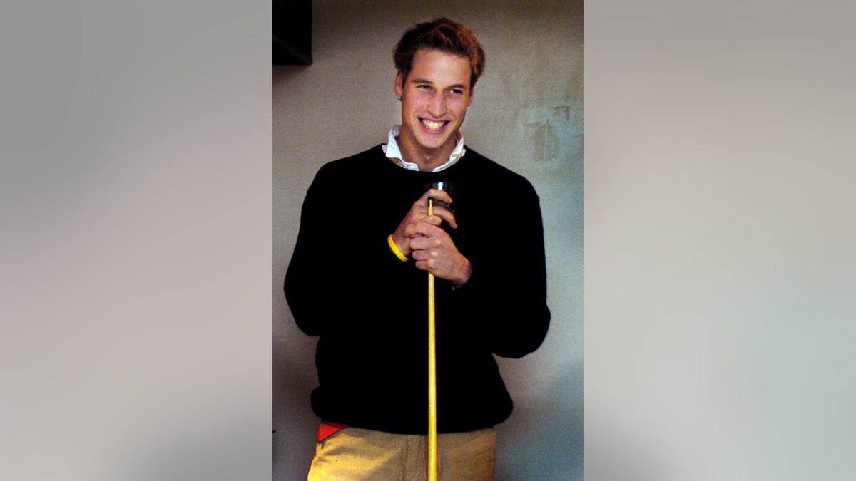 Prince William holding a billiards cue wearing a black sweater, a white collared shirt and pants.