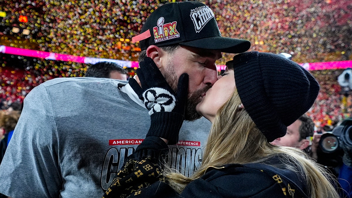 Travis Kelce wearing a grey AFC Champions t-shirt and hat kisses Taylor Swift in a black hat and gloves while confetti falls down on the field