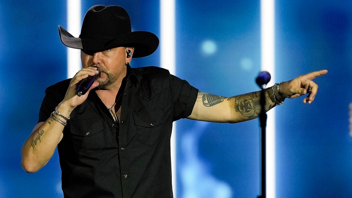 Jason Aldean wearing a black short-sleeved shirt and black cowboy hat performing on stage, extending his left arm at the Liberty Ball