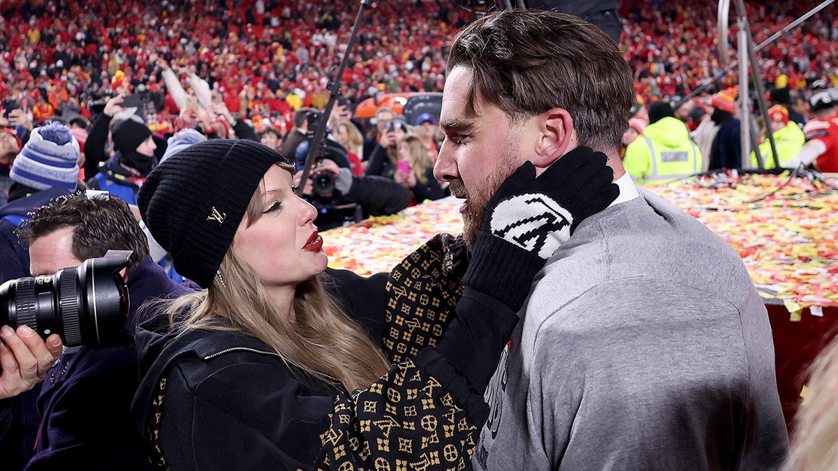 Tatkir Swift in a black LV beanie looks up as she grabs Travis Kelce's face wearing black LV mittens