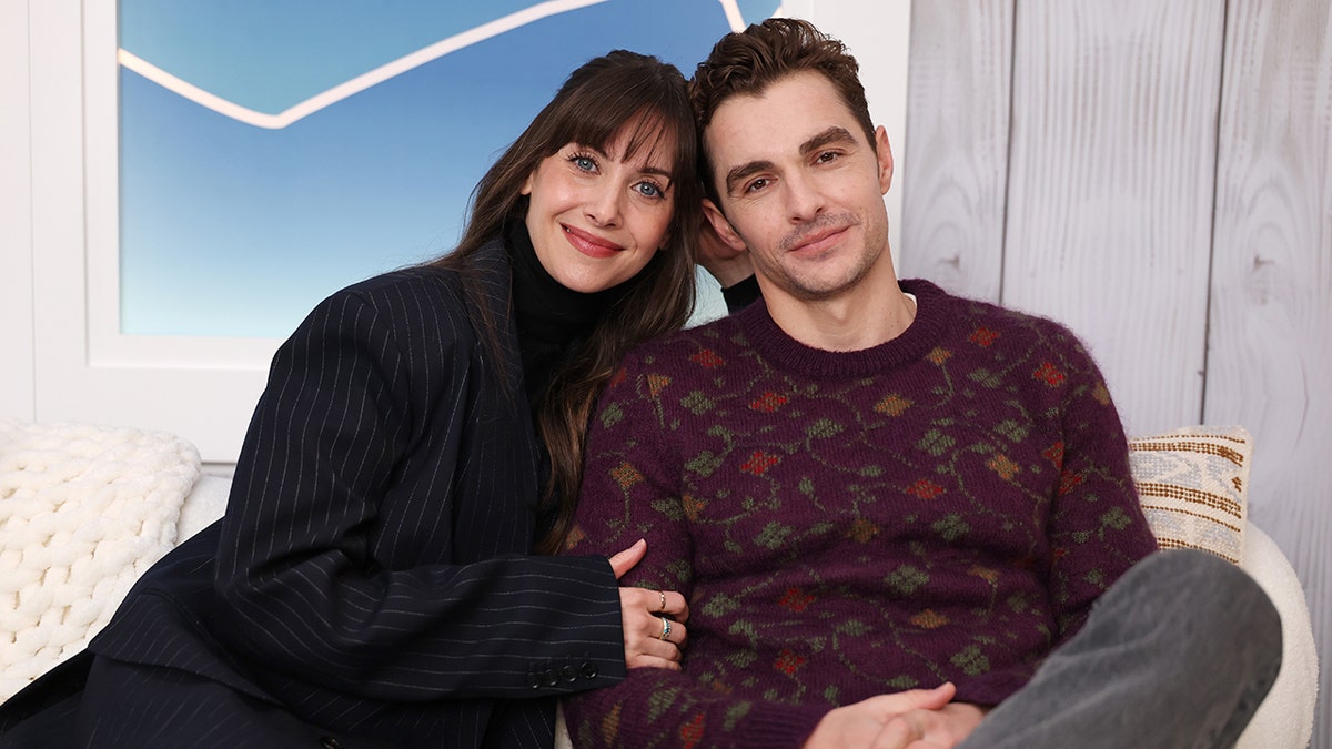 Alison Brie soft smiles wearing a black striped suit holding on to husband Dave Franco wearing a deep purple floral sweater