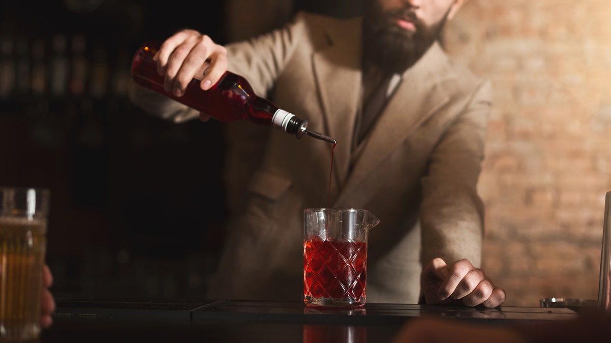 A bartender pouring red liquor or grenadine syrup into a glass, preparing alcoholic cocktails at a bar counter.