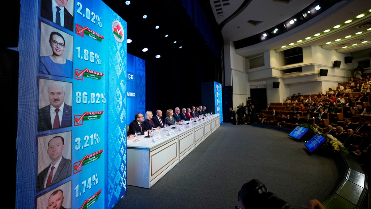 The results of Belarus's 2024 elections are displayed on the screens next to the long table where the Belarusian bureaucrats are sitting.