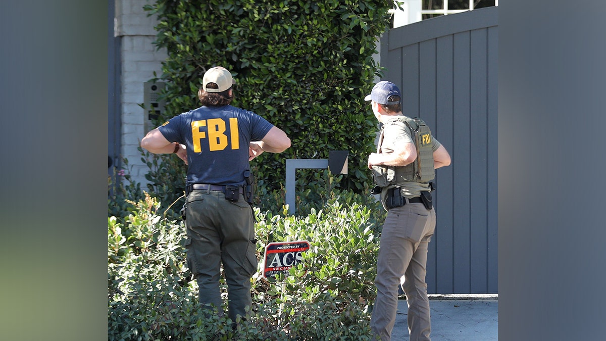 Outside Ben Affleck's home, a man wearing a navy blue FBI jacket stands with his back to the camera next to another gentleman wearing a green FBI jacket