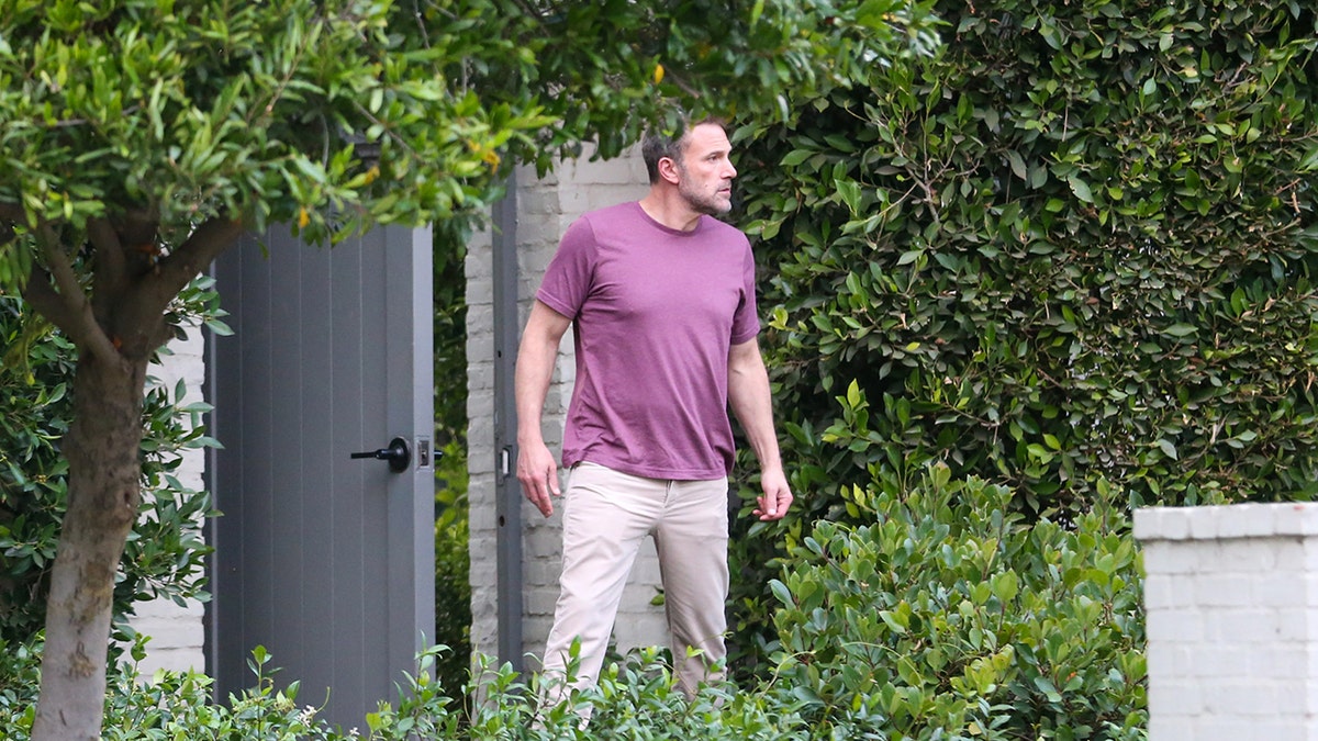 Ben Affleck in a maroon shirt and tan slacks looks angsty as he stands outside his home in Brentwood