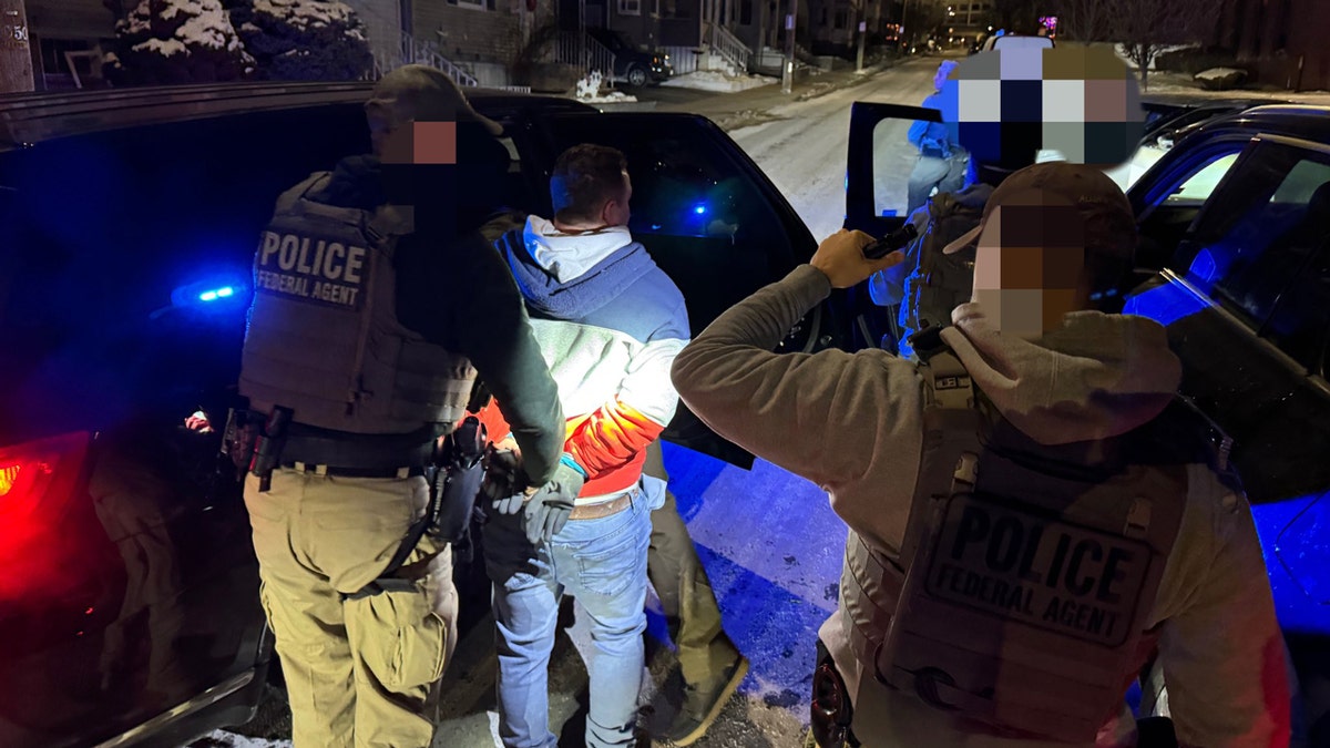 ICE officers lead a man in handcuffs into a police car.