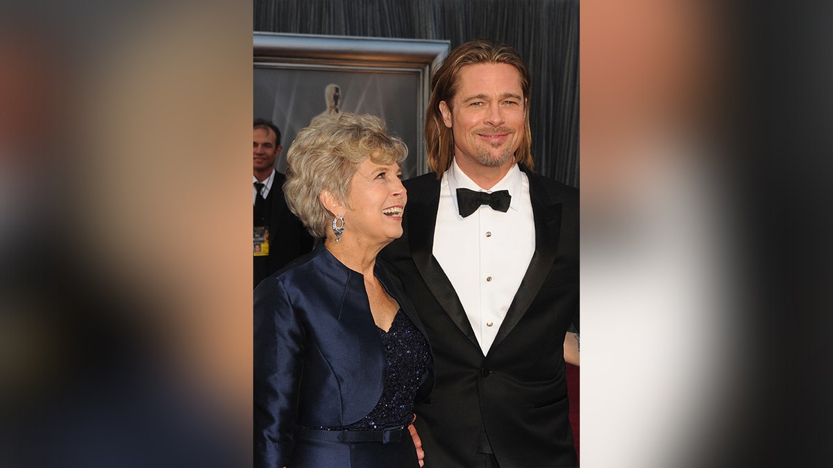 Jane Pitt in a navy blue gown with a sparkly bust and overcoat looks upu and smiles at son Brad Pitt in a classic tuxedo at the Academy Awards