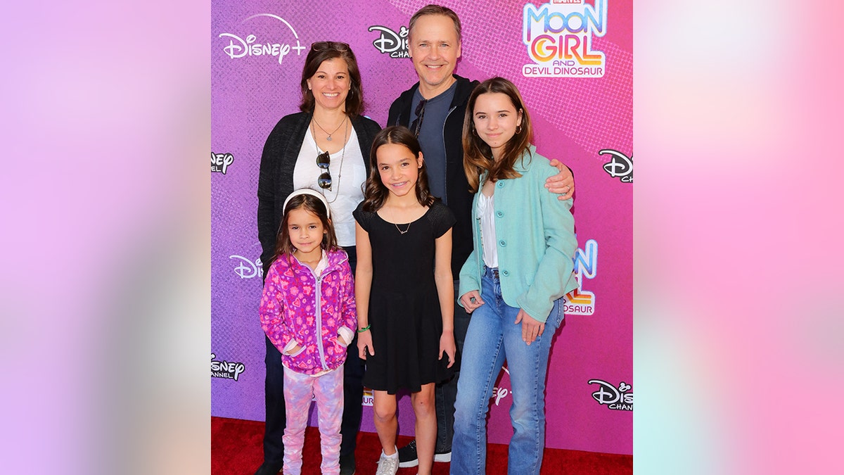 Chad Lowe on the carpet in a black sweatshirt wraps his arms around one of his three young daughters, also standing next to his wife Kim in a black cardigan and white t-shirt