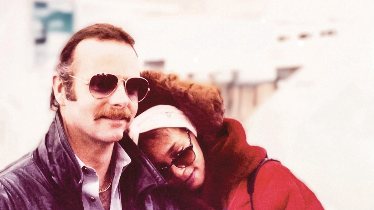 David Roberts wearing sunglasses and a dark jacket with a plaid shirt next to Whitney Houston in a red coat and glasses with her head down.