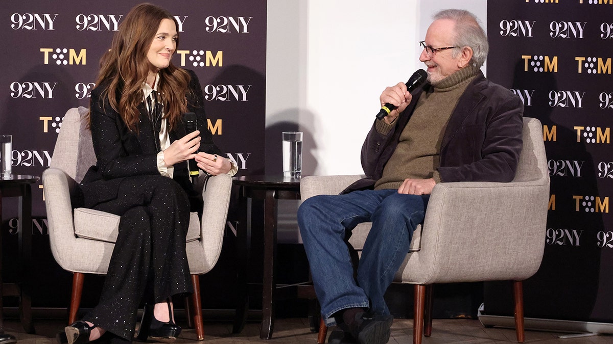 Drew Barrymore in a sparkly black pantsuit smiles on stage at Steve Spielberg sitting opposite her in blue jeans and a velvet brown suit