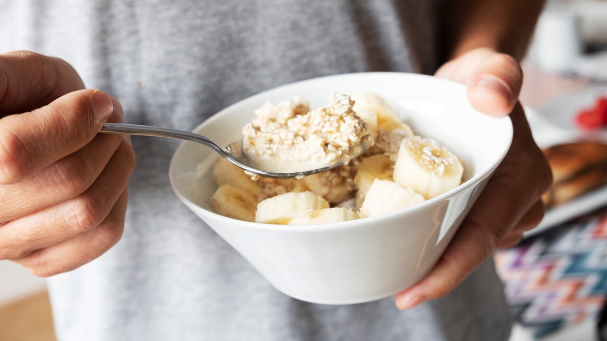 A man is holding a bowl of oatmeal with bananas in one hand and a spoon in the other.