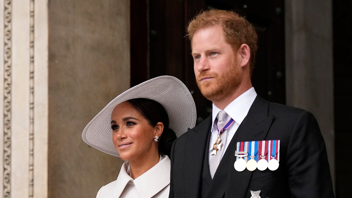 Prince Harry wears a suit with medals and stands next to Meghan Markle in a white coat with matching hat.