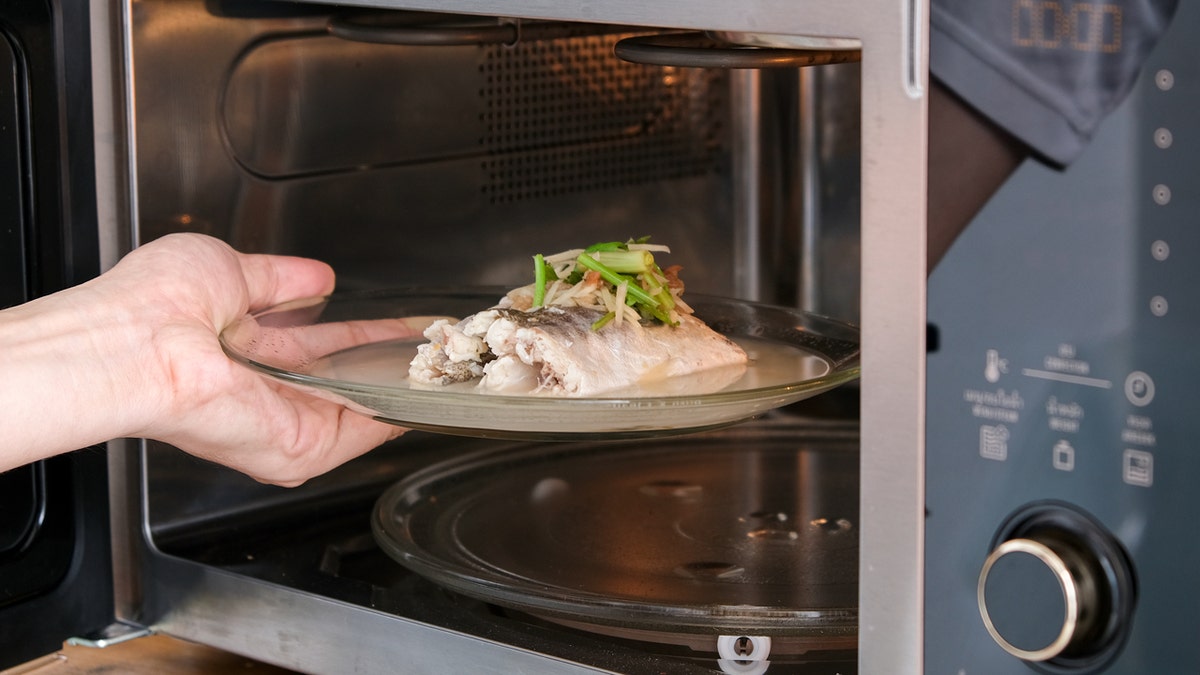 A hand puts a plate of fish in the microwave.