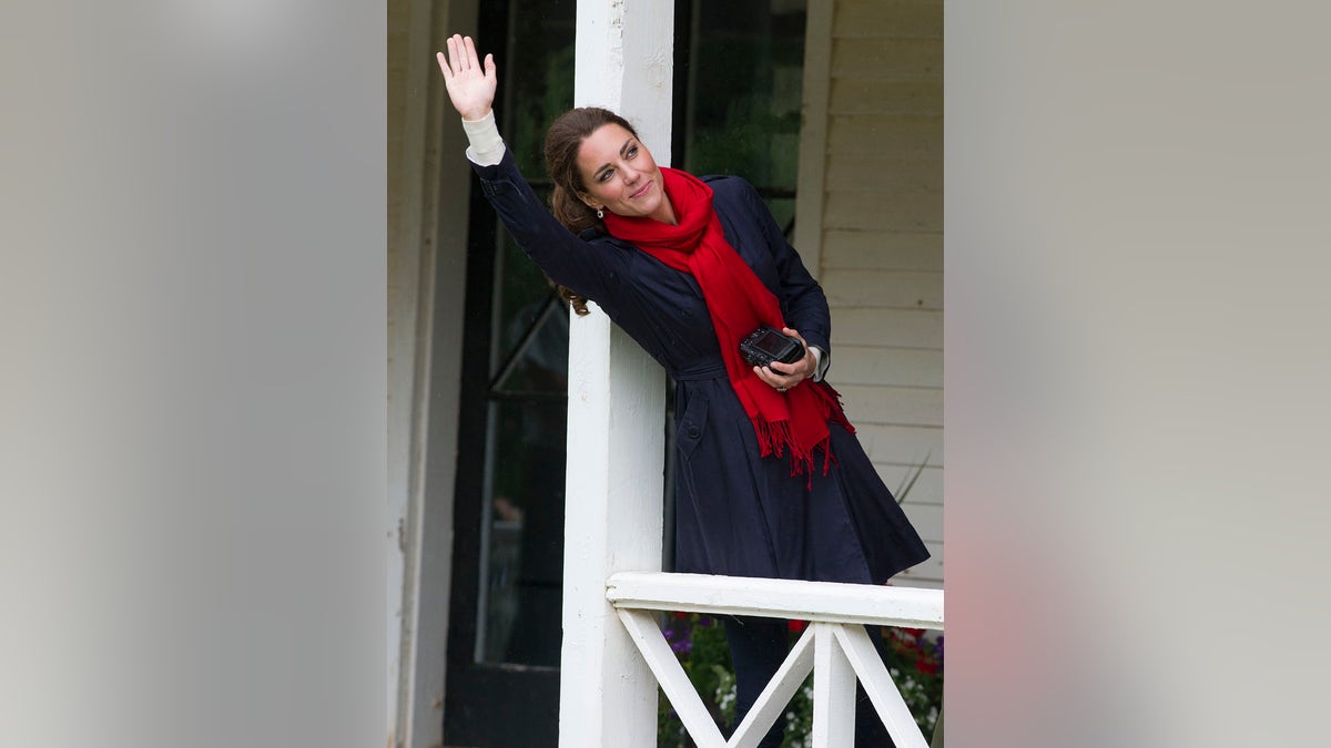 Kate Middleton holding a camera while wearing a blue coat and a red scarf waving