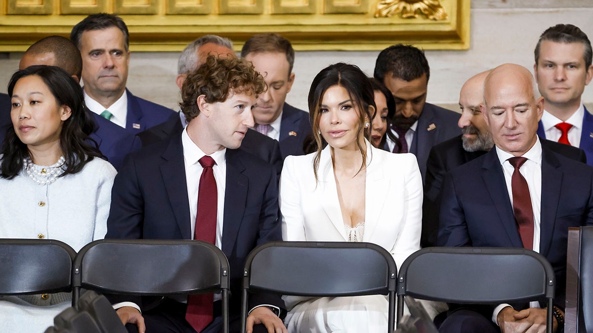 Lauren Sanchez in a white suit sitting in between Mark Zuckberg and Jeff Bezos.