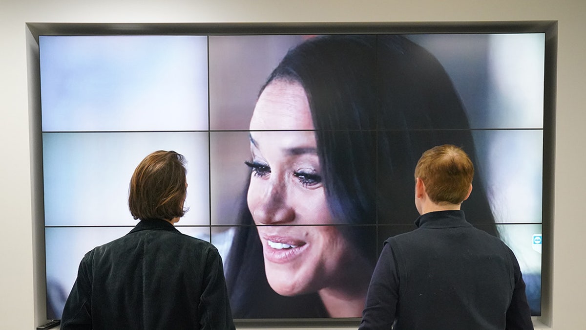 Two people with their backs turned watching Meghan Markle on television.
