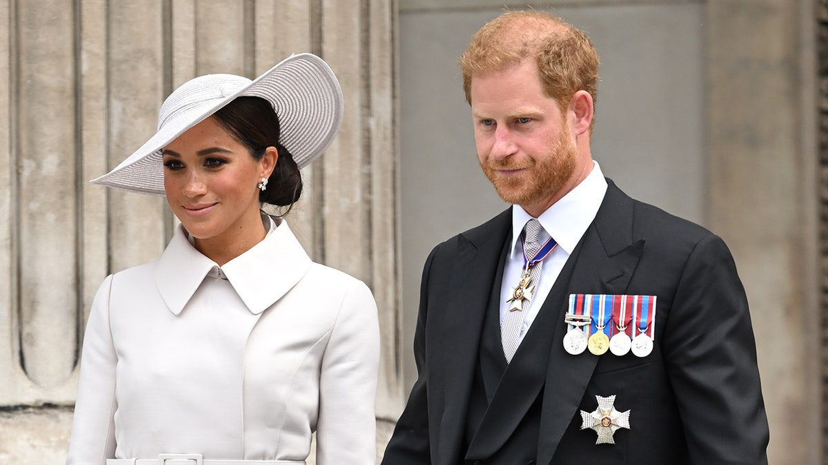 Meghan Markle con un abrigo blanco y un sombrero a juego caminando junto al Príncipe Harry con un traje con medallas.