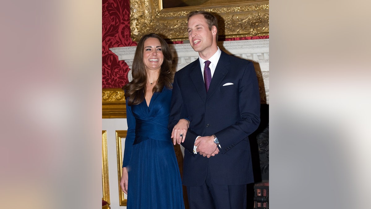 Kate Middleton smiling wearing a royal blue wrap dress in Prince William's arm. He is smiling wearing a navy suit and a purple tie.