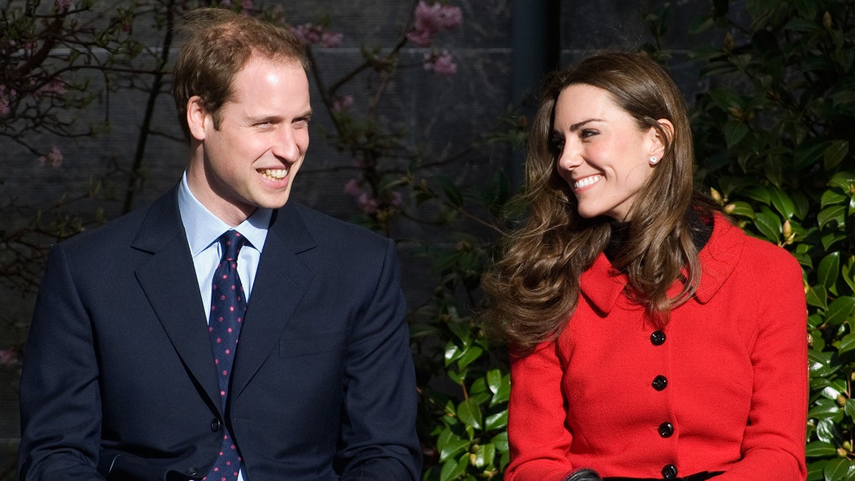 Prince William wearing a dark blue suit smiling at Kate Middleton wearing a bright red coatdress