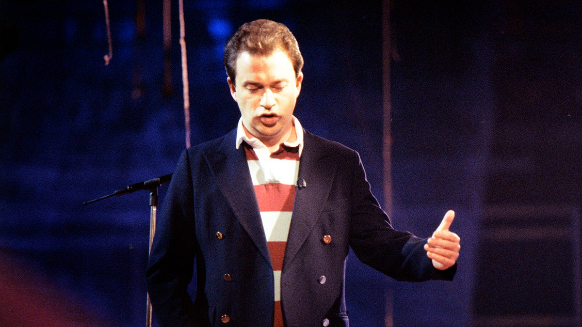 Harry Enfield acting out a scene as Tim Nice But Dim wearing a navy coat and a red and white striped shirt on stage.