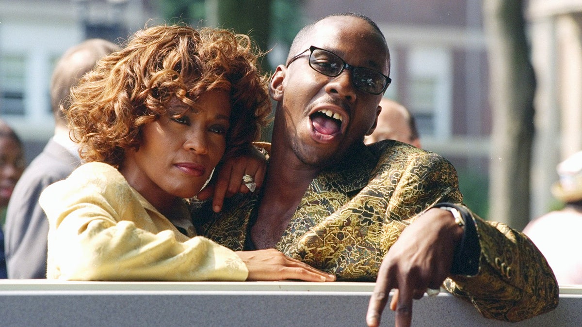 Bobbi Brown in a gold blazer shouting with Whitney Houston wearing a yellow sweater with her arms around him.