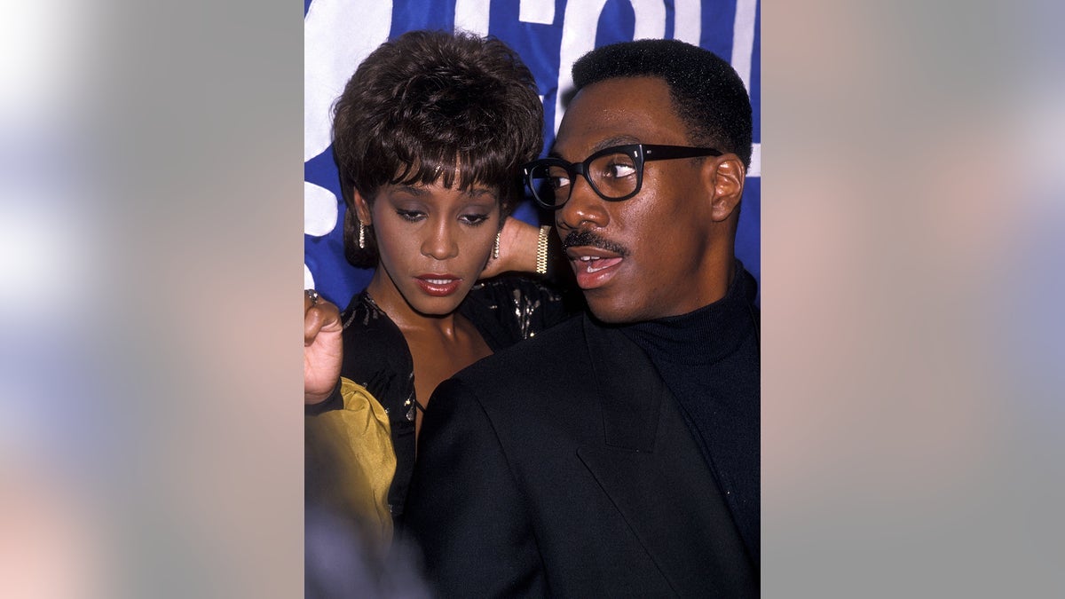 Whitney Houston looking down wearing a gold and black blazer as Eddie Murphy, wearing all black, looks away.