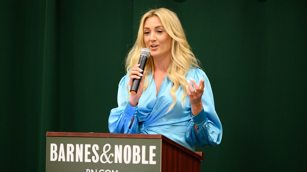 Liana Werner-Gray speaking at a dais. She is wearing a blue top and she is holding a microphone.