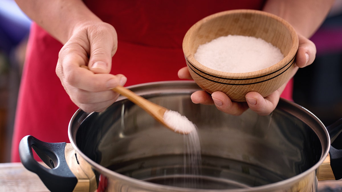 Pasta cooking - hand holding wooden bowl with salt, seasoning water with sea salt.