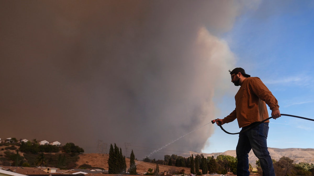 Andrew Aguilar sprays water from the top of the roof at his brother's home Castaic, Calif., as a large plume of smoke caused by the Hughes Fire rises from Castaic Lake on Wednesday.