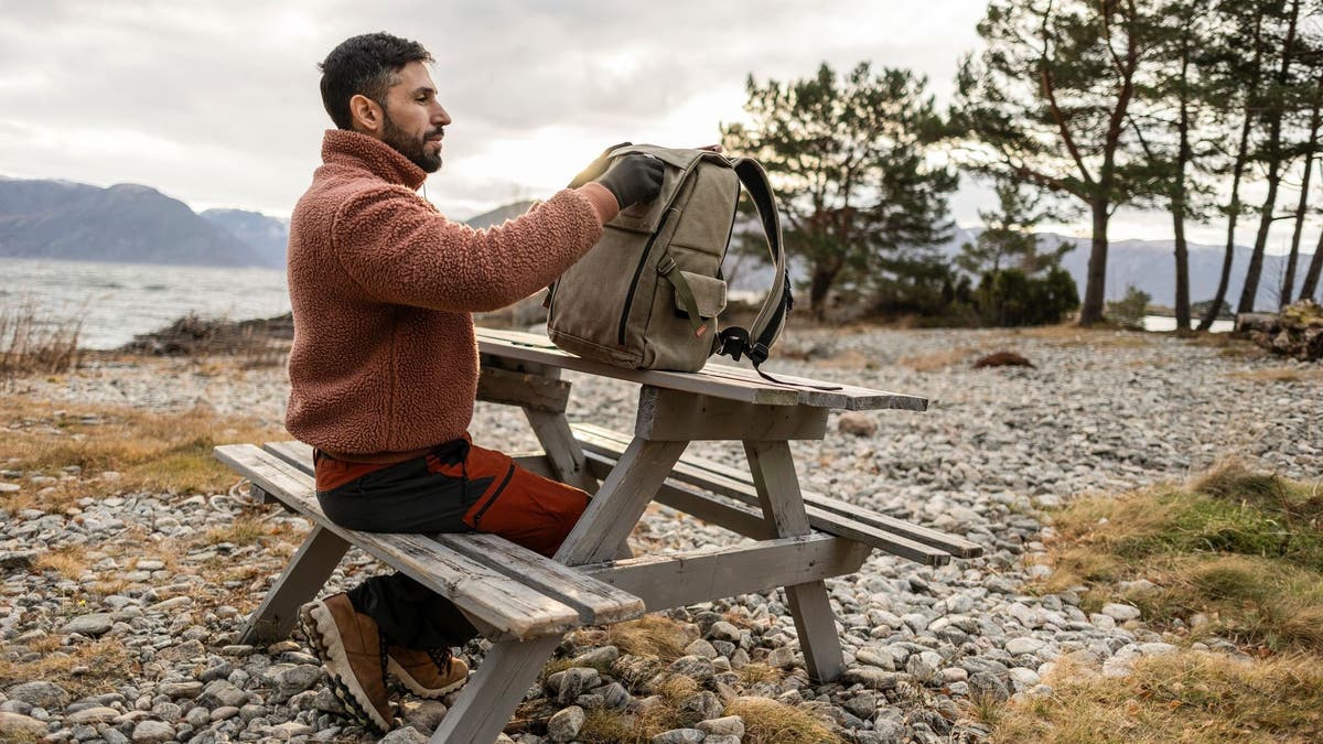 A man in a cozy sweater packs his bag at a picnic table, with a calm lake and mountains in the distance.