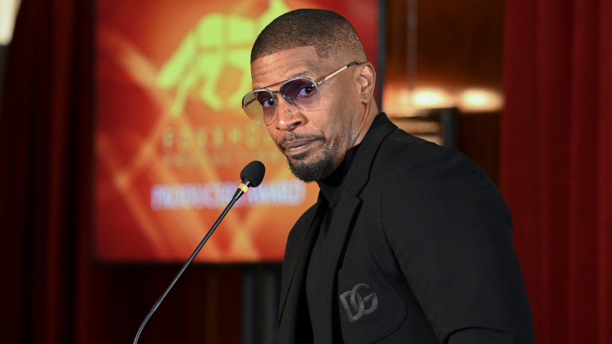 Jamie Foxx in a black Dolce & Gabbana suit and transparent lens sunglasses makes a silly face standing behind the podium