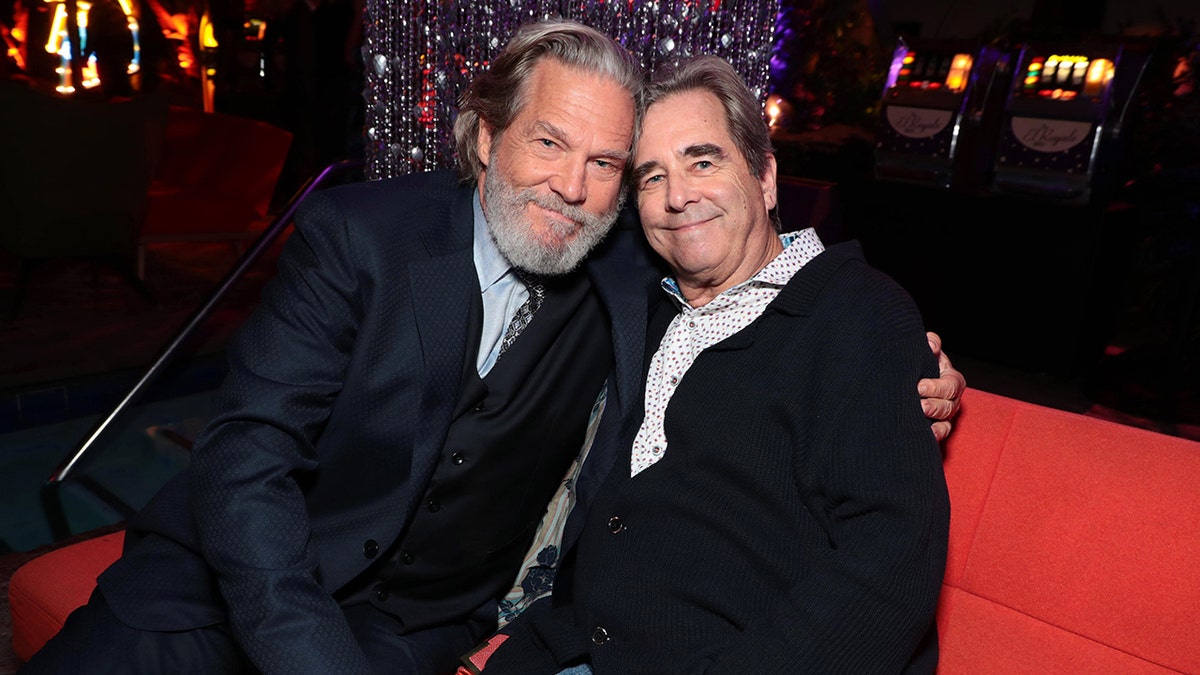 Jeff Bridges in a navy jacket leans in on a couch and hugs his brother Beau in a black cardigan as they both soft smile for a picture