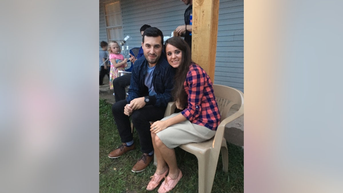 Jinger Duggar Vuolo dengan kemeja kotak-kotak biru dan merah muda duduk di kursi krem ​​​​dan bersandar ke arah suaminya Jeremy Vuolo dengan setelan jas biru.