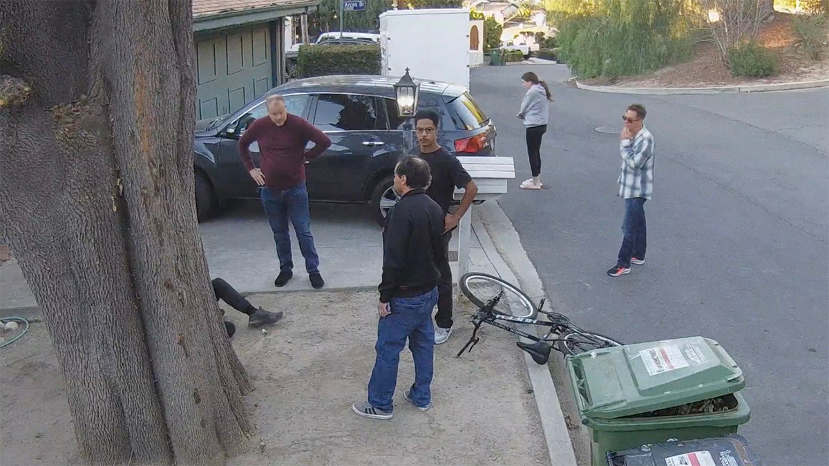 Actor Brian J. White stands with his hands on his hips outside his neighbors house surrounded by neighbors while a man sits on the ground behind a tree