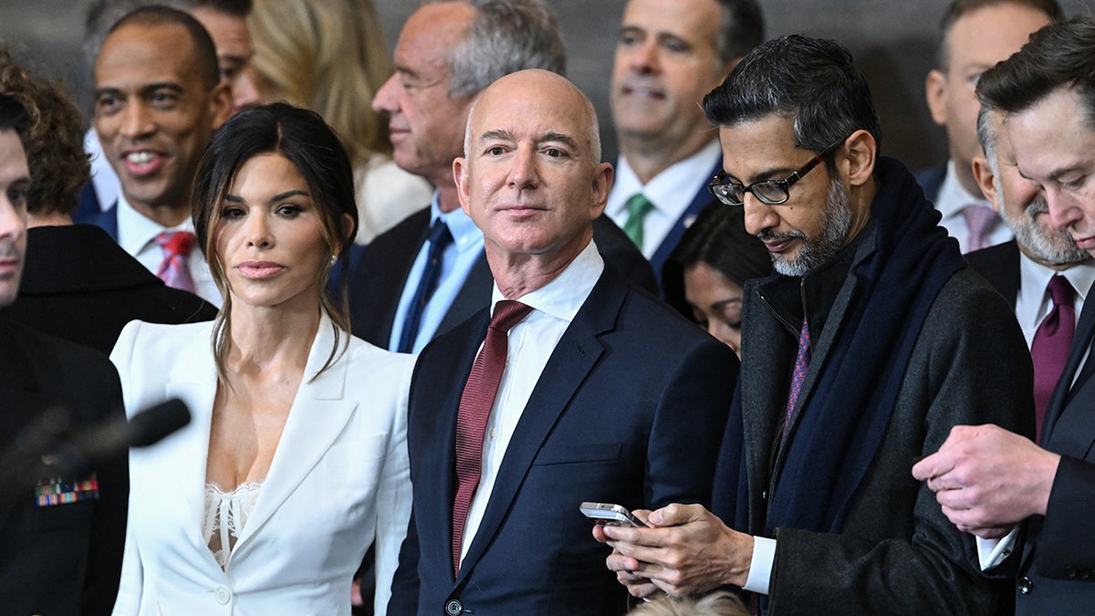 Lauren Sánchez in a white blazer and busty lace white bra/top stands beside Jeff Bezos in a dark suit and dark red tie at the inauguration