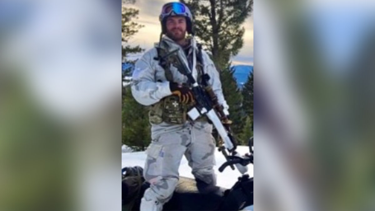 Matthew Livelsberger wears cold weather gear and carries a rifle while standing on top of a snowmobile