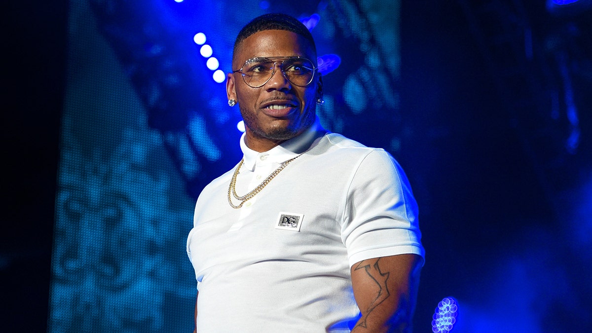 Nelly is wearing a white polo shirt and chain necklace with clear glasses and looks to his left on stage