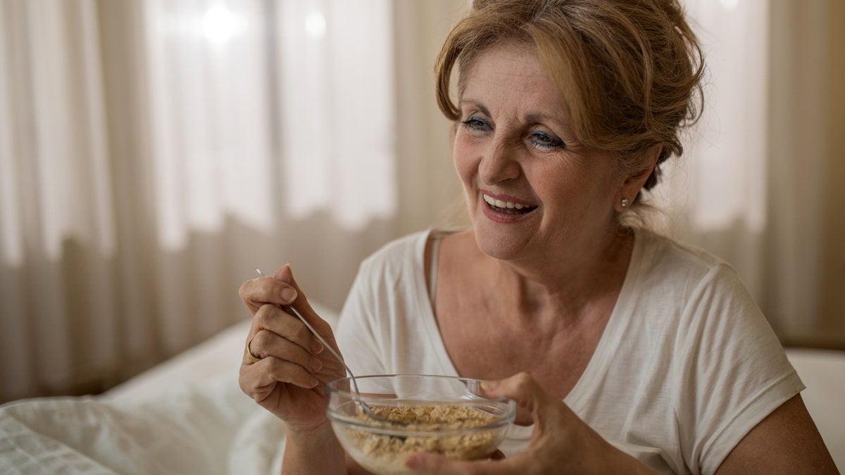 Mature woman eating oatmeal in bed smiling.