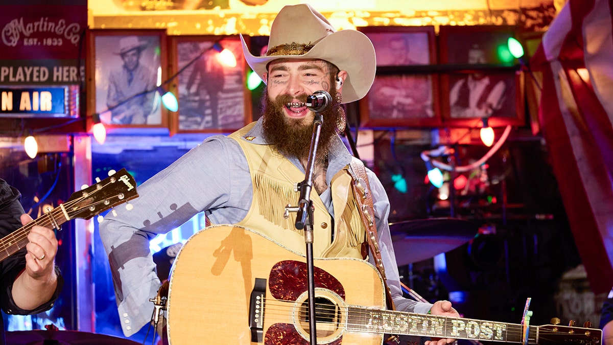 Post Malone dengan rompi kuning dan kemeja biru tersenyum saat berada di atas panggung dengan topi koboi berwarna coklat