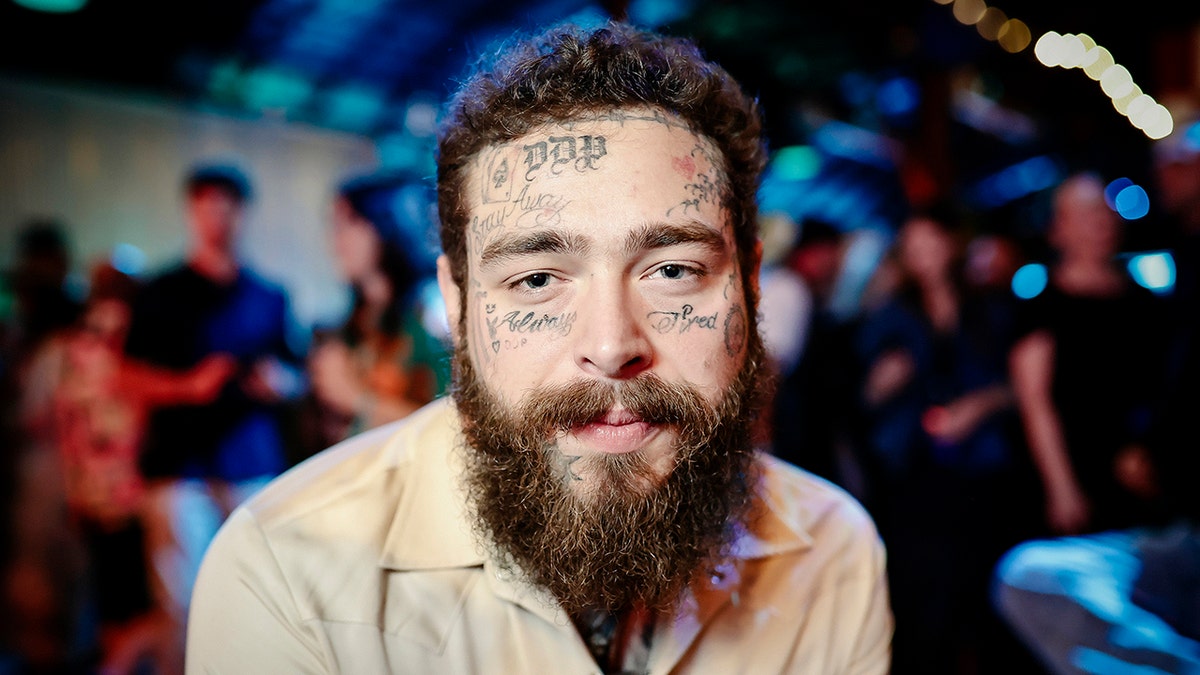 Post Malone looks directly at the camera wearing a tan shirt