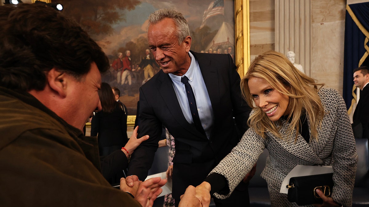 Robert F. Kennedy Jr. in a black suit and tie and wife and actress Cheryl Hines in a houndtooth jacket lean over and shake the hands of people as they leave the inauguration