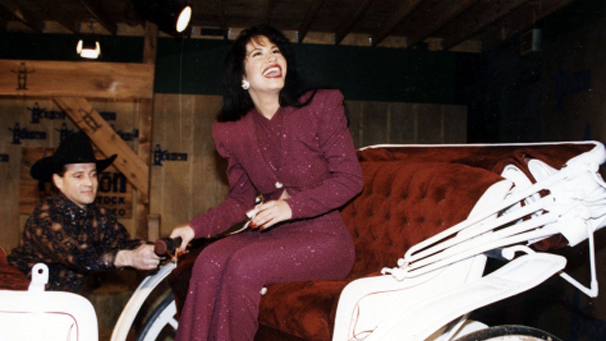 Selena Quintanilla in a maroon pantsuit looks up and laughs as she sits in a horse-drawn carriage