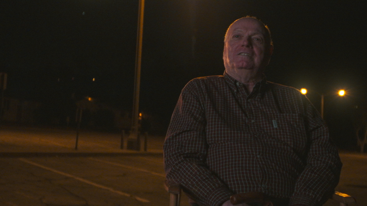 Skip Junis is seated in a char outdoors speaking with an interviewer at nighttime