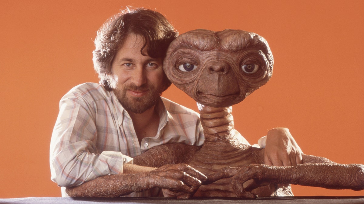 Steven Spielberg in plaid shirt smiling with ET in front of orange background drop