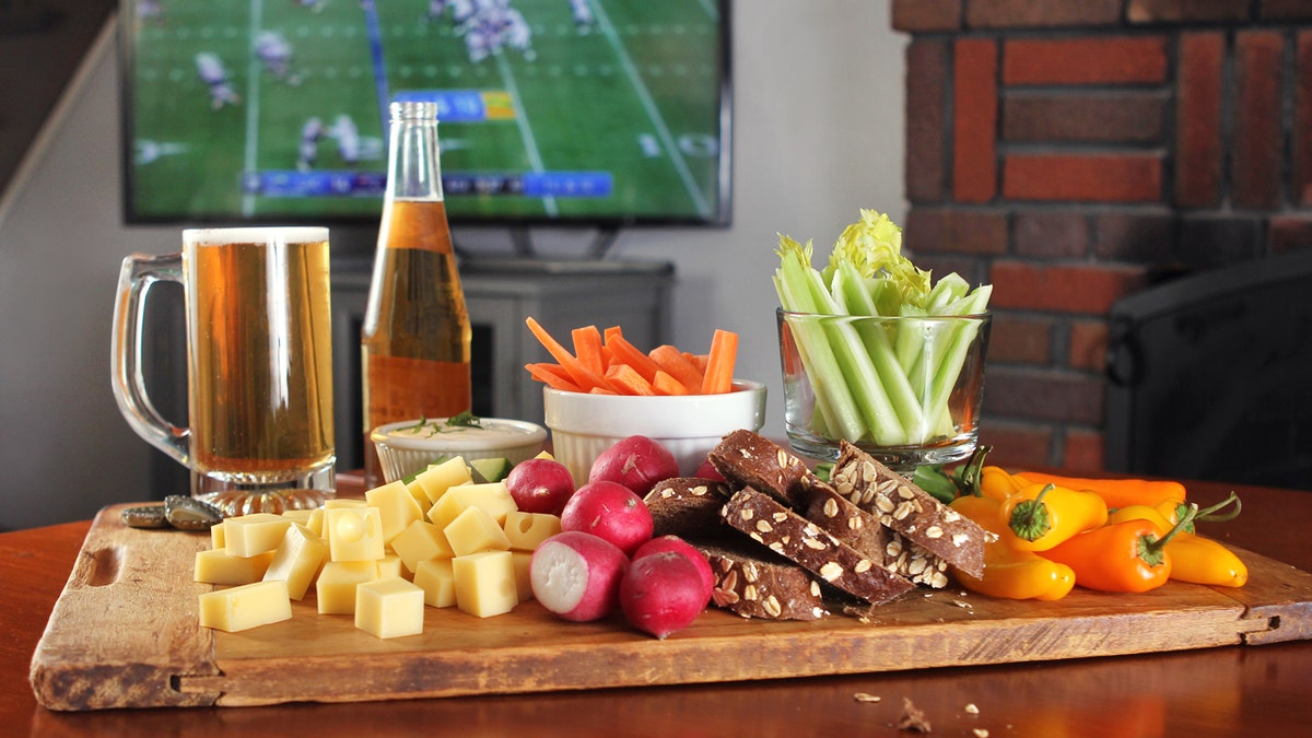 Football game plays on TV in the background. In the foreground, there are healthy food options for parties and some beer.