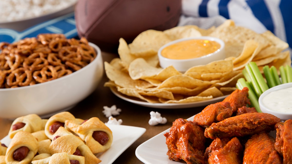 Super Bowl party foods include, clockwise starting from top right, chips and queso, chicken wings with celery, pigs in a blanket and pretzels.