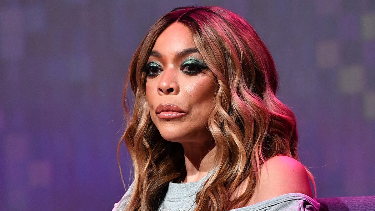 Wendy Williams con una camisa gris con un hombro recortado mira hacia abajo y abatida en el escenario