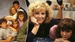 ABC Photo Archives/Disney General Entertainment Content via Getty Images split Joanna Kerns as Maggie Seaver puts her hand on her chin as she lies on the bed next to Tracey Gold in a pink sweater and her arm underneath her neck
