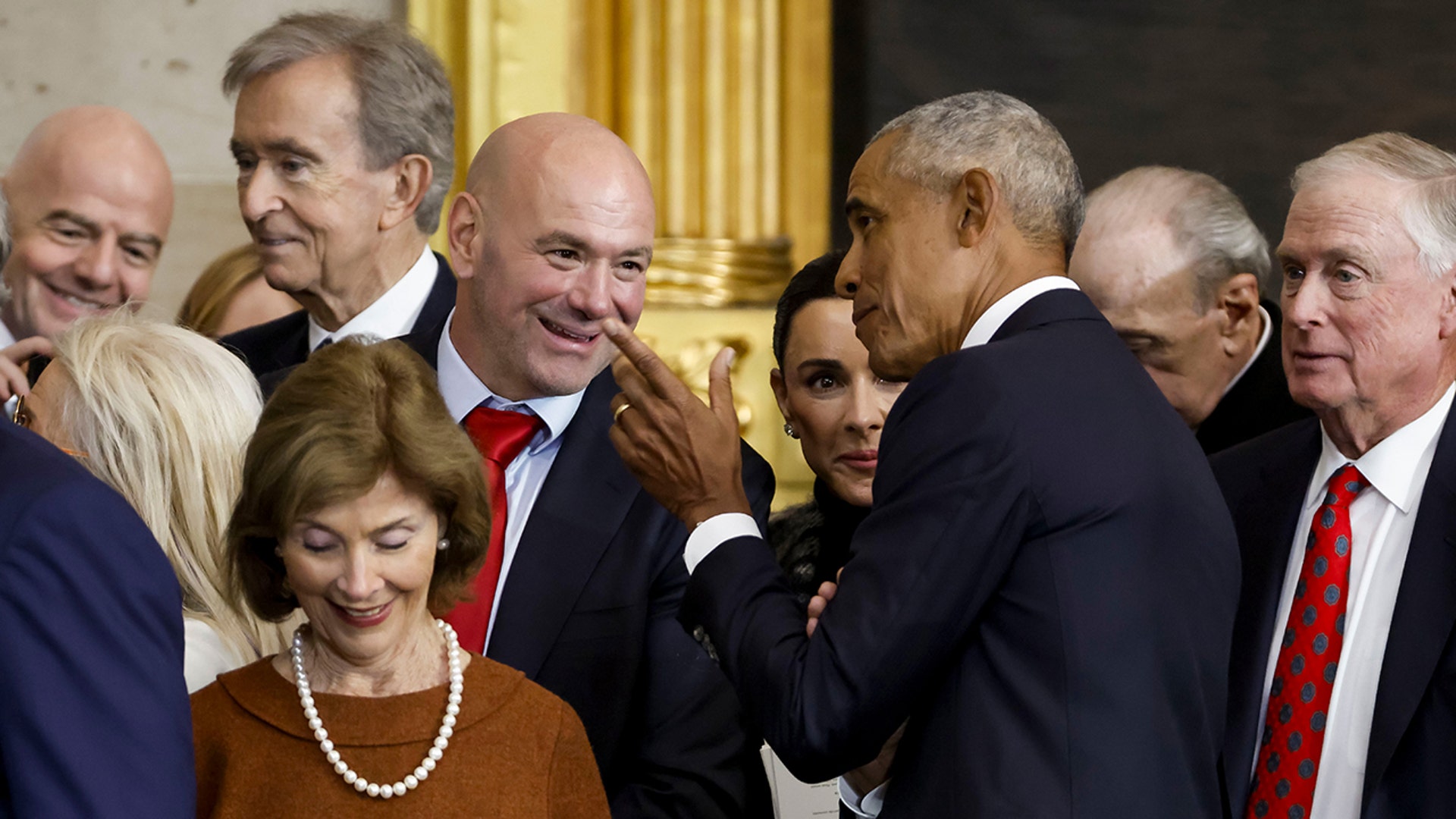 Dana White in a dark suit and red tie converses with former President Obama as his wife stands beside him and former First Lady Laura Bush walks out of frame